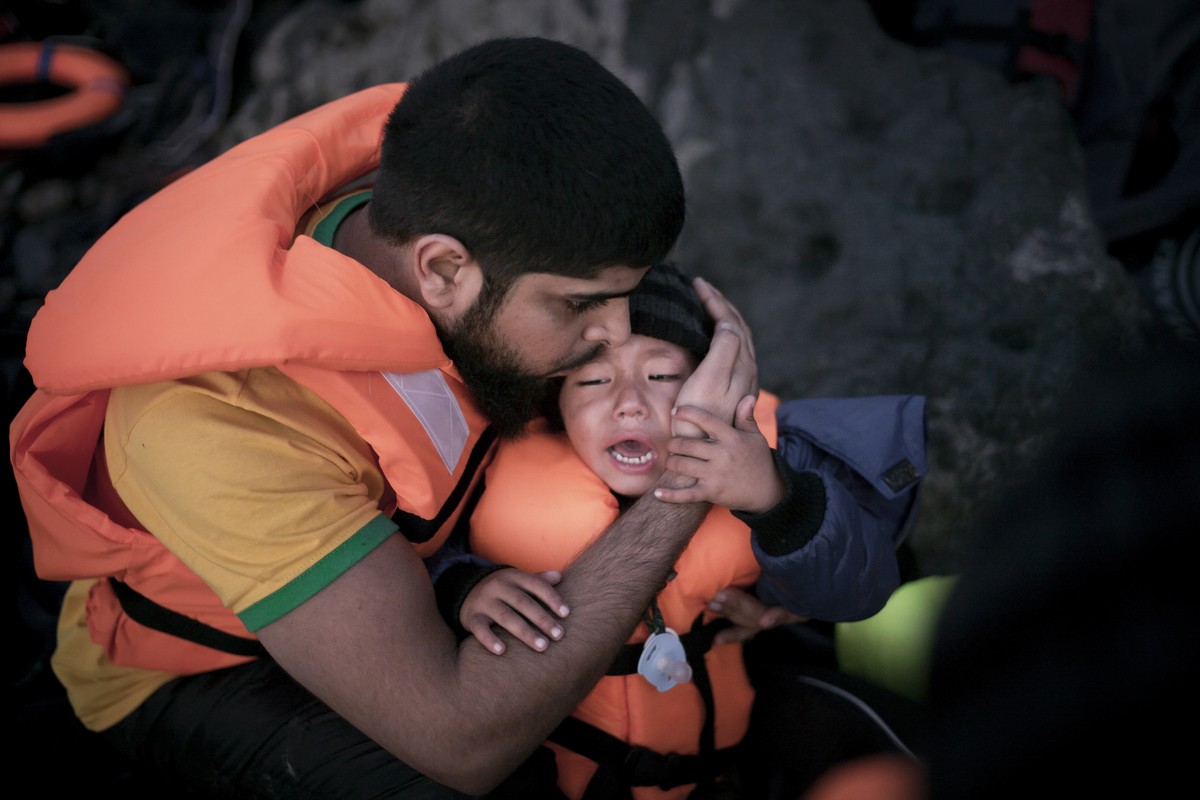 En far kramar om sin gråtande son. Båda är iförda flytvästar och en livräddningsboj ligger en bit bort.