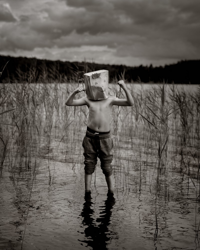 Ett barn står i vattenbrynet i en sjö med gles vass och skog runt. Barnet har en papperspåse på huvudet med ett ritat ansikte. Armarna är lyfta och spända som för att visa sina muskler på den bara överkroppen.