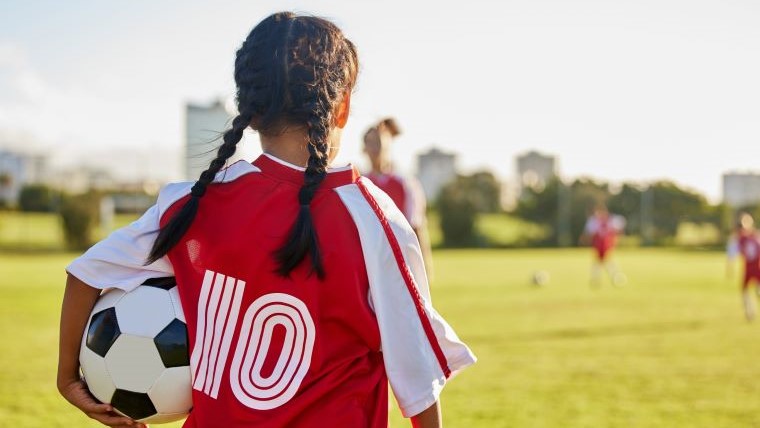 Ett barn står på en fotbollsplan med en fotboll under armen. 